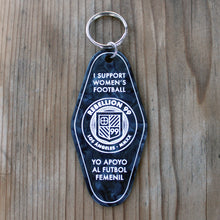 Load image into Gallery viewer, Front side of Rebellion 99 keychain is shown against a wooden background. The black acrylic engraved keychain is shown and details are filled with white enamel to form the design. A silver keychain is attached at the top of each keychain. Front side reads &quot;I support women&#39;s football&quot; and &quot;Yo apoyo al futbol femenil&quot; with the Rebellion 99 crest in the middle.

