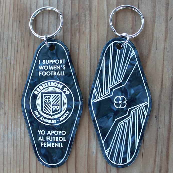 Both front and back side of Rebellion 99 keychain are shown against a wooden background. The black acrylic engraved keychain is shown and details are filled with white enamel to form the design. A silver keychain is attached at the top of each keychain.