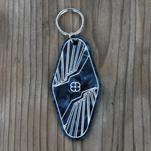 Load image into Gallery viewer, Back side of Rebellion 99 keychain is shown against a wooden background. The black acrylic engraved keychain is shown and details are filled with white enamel to form the design. A silver keychain is attached at the top of each keychain. Back side shows Rebellion 99 poppy and wing design.
