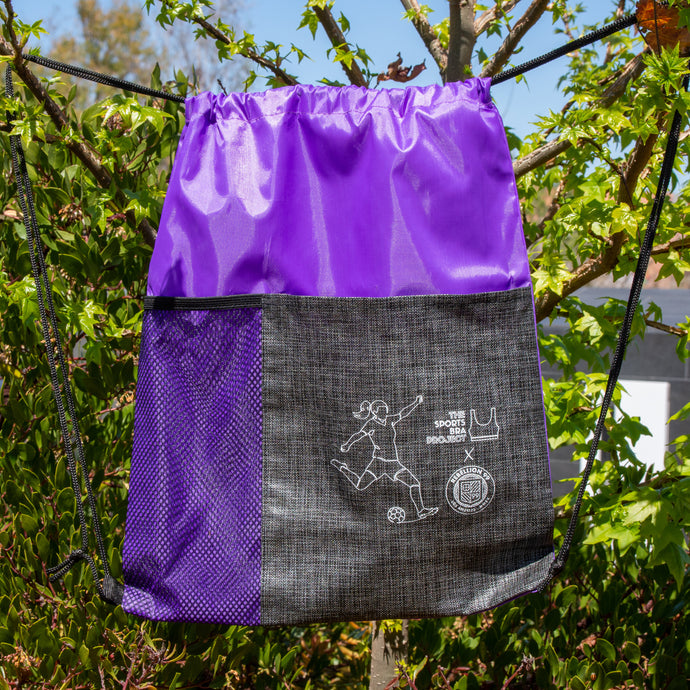 Purple drawstring backpack is shown in front of green trees.